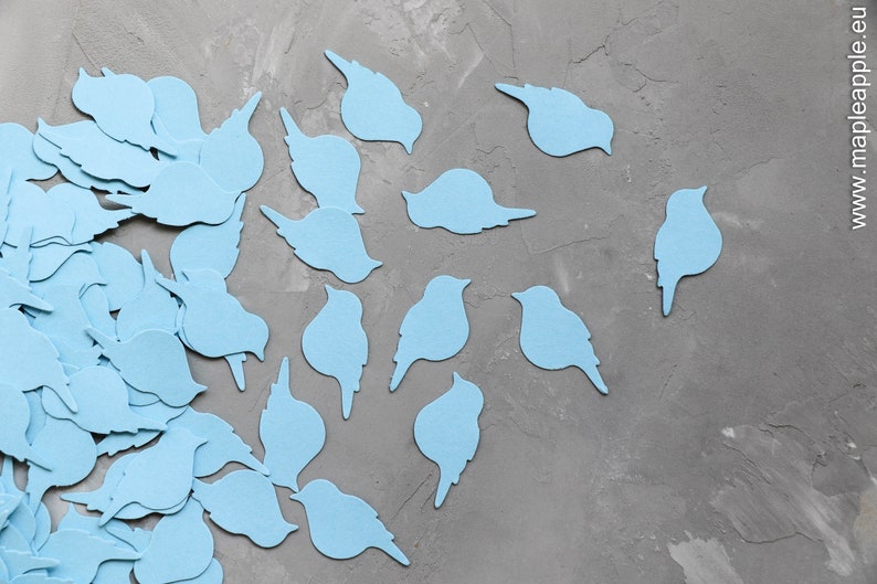 May include: Light blue bird-shaped cutouts scattered on a grey textured surface. The cutouts vary in shape, some with wings and others without, creating a whimsical and decorative effect. The image suggests a craft or DIY project.