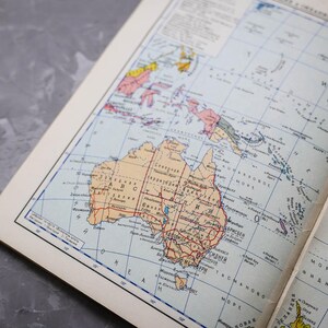 May include: Open book with a vintage map of Australia and surrounding islands. The map is in shades of tan, blue, and red, with detailed geographical features and text. The book's pages are off-white, and the background is a gray surface.