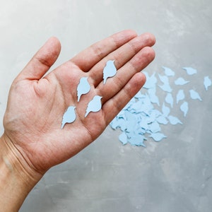 May include: A hand displaying several light blue bird-shaped paper cutouts, with more scattered on a grey surface. The birds are uniformly coloured and appear to be decorative elements, possibly for crafts or event decorations.