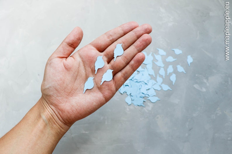 May include: A hand displaying several light blue bird-shaped paper cutouts, with more scattered on a grey surface. The birds are uniformly coloured and appear to be decorative elements, possibly for crafts or event decorations.