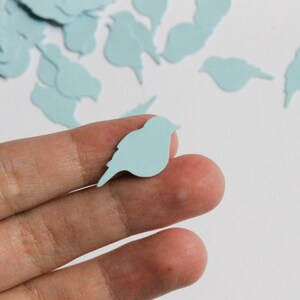 May include: A hand holds a light blue bird-shaped confetti piece, with more scattered in the background. The confetti is a pale blue, and the bird shape is a simple silhouette. The image suggests a craft or decorative theme.