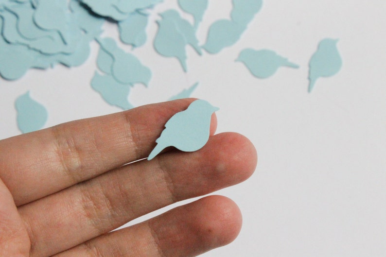 May include: A hand holds a light blue bird-shaped confetti piece, with more scattered in the background. The confetti is a pale blue, and the bird shape is a simple silhouette. The image suggests a craft or decorative theme.