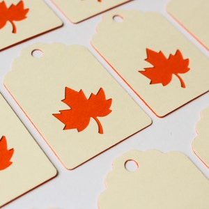 May include: Cream-colored gift tags with a die-cut orange maple leaf design. The tags have a scalloped edge and a small hole at the top for stringing. The image shows multiple tags, perfect for fall-themed crafts or gifts.