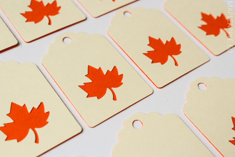 May include: Cream-colored gift tags with a die-cut orange maple leaf design. The tags have a scalloped edge and a small hole at the top for stringing. The image shows multiple tags, perfect for fall-themed crafts or gifts.