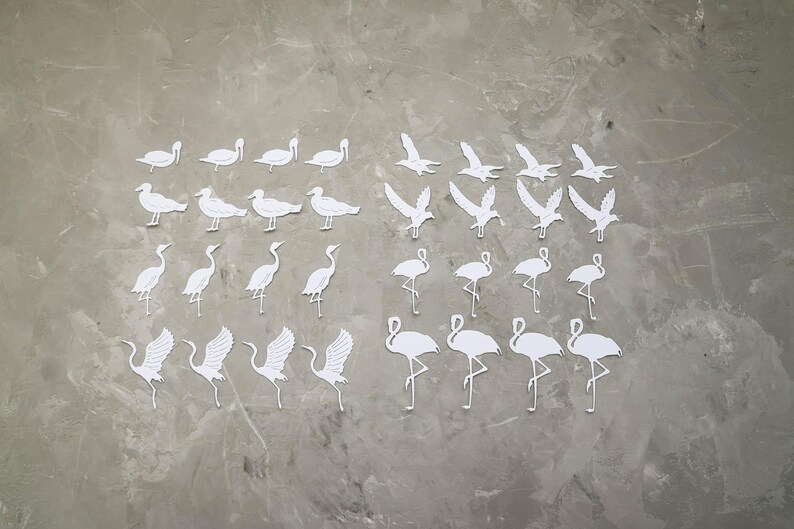 May include: A selection of white paper bird cutouts, including swans, seagulls, cranes, and flamingos, displayed on a grey surface. The birds are depicted in various poses, some with wings extended, others standing or in flight.