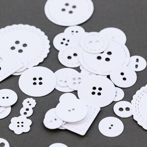 Puede incluir: Una colección de botones de papel blanco de varios tamaños y formas, incluyendo bordes redondos, cuadrados y festoneados. Cada botón tiene dos o cuatro agujeros, algunos con costuras decorativas. Los botones están dispersos sobre una superficie gris oscuro.