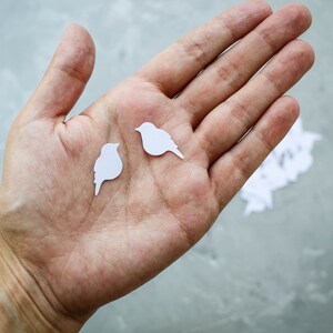 May include: Two small, white paper bird cutouts are held in an open hand. The birds have simple, stylized shapes with a beak and tail. The background is a mottled grey surface. The website address www.mapleapple.eu is visible in the upper right corner.