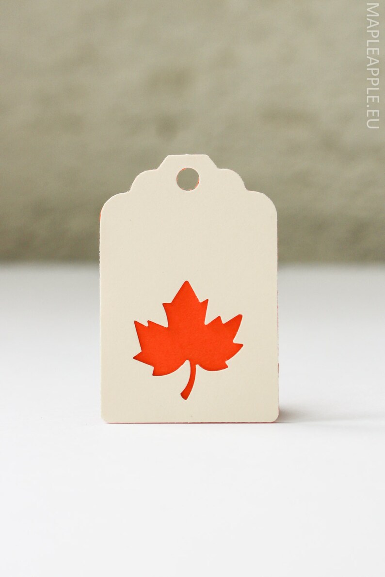 May include: Cream-colored tag with a die-cut orange maple leaf design. The tag has a decorative top edge and a hole for hanging. The website mapleapple.eu is printed on the right side.