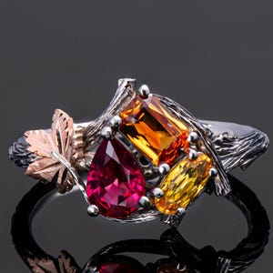 Könnte beinhalten: Ein silberner Ring mit farbigen Edelsteinen. Er hat einen tropfenförmigen Rubin, einen rechteckigen orangefarbenen Stein und einen gelben ovalen Stein. Ein Design aus einer roségoldenen Blattform und silbernen Zweigen rundet den Ring ab.