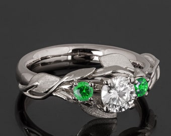 Könnte beinhalten: Ein silberner Ring mit einem weißen Diamanten und zwei grünen Edelsteinen, die in einem Blattdesign gefasst sind.