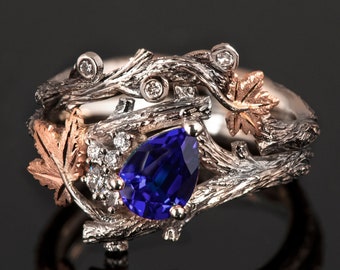 Puede incluir: Un anillo de plata y oro rosa con una piedra de zafiro azul en forma de pera engastada en un diseño de rama con pequeños diamantes y hojas.