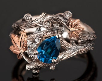 Puede incluir: Un anillo de plata y oro rosa con una piedra preciosa azul en forma de pera engastada en un diseño de rama con pequeños diamantes y hojas.