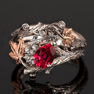 Könnte beinhalten: Ein Ring aus Silber und Roségold mit einem birnenförmigen Rubin, der von kleineren Diamanten umgeben ist. Der Ring ist mit ineinander verschlungenen Ästen und Blättern verziert.