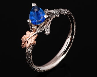Puede incluir: Un anillo de plata con una piedra preciosa azul engastada en un diseño de rama. El anillo presenta una pequeña hoja de oro rosa.