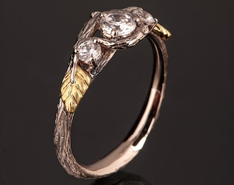 May include: A delicate gold and silver ring with a twig-like band and three round diamonds. The band is textured to resemble bark and has a small gold leaf detail.
