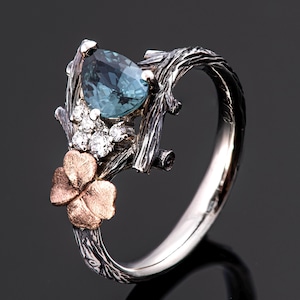 Peut inclure: Bague en argent ornée d'une pierre précieuse bleu clair en forme de poire et de petits diamants blancs. La bague présente un motif en forme de branche avec un trèfle à quatre feuilles en or rose.