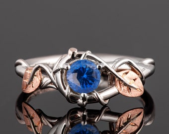 Puede incluir: Un anillo de plata y oro rosa con una piedra preciosa de zafiro azul engastada en un diseño de vid. El anillo presenta dos hojas, una a cada lado de la piedra.