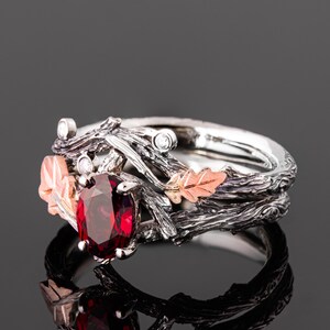 Op de afbeelding: Een zilveren en roségouden ring met een rode edelsteen gezet in een takvormig ontwerp. De ring is versierd met kleine diamanten en bladeren.