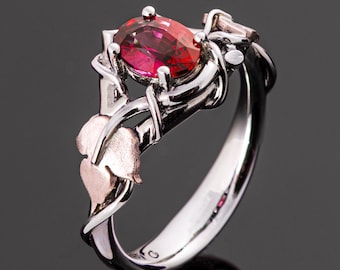 May include: A silver ring with an oval, red gemstone. The ring has a floral design with rose gold accents on the leaves. The band is polished and smooth, with intricate detailing around the gemstone.