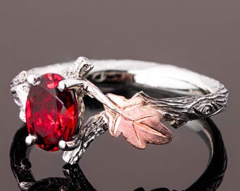 May include: A silver ring with a red gemstone set in a branch-like design. The ring features a rose gold leaf on one side.