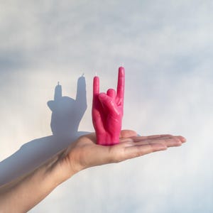 Könnte beinhalten: Eine leuchtend pinke Kerze in Form einer Hand, die das Rock-and-Roll-Zeichen formt. Die Kerze hat zwei weiße Dochte und wirft einen Schatten auf einen weißen Hintergrund. Die Kerze wird in der Hand einer Person gehalten.