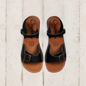 May include: A pair of black leather sandals with a buckle closure. The sandals have a brown leather sole and are made by the brand "Crupon".