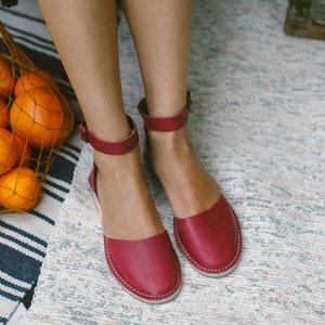 Könnte beinhalten: Ein Paar rote Ledersandalen mit Knöchelriemen. Die Sandalen befinden sich auf einem weiß-blau gestreiften Teppich.