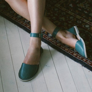 Könnte beinhalten: Eine Frau trägt einen gestreiften Jumpsuit mit einem braunen Bandana um den Hals. Sie sitzt auf einem Teppich mit gekreuzten Beinen und trägt türkisfarbene Knöchelriemensandalen.