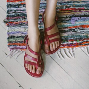 May include: A pair of red leather sandals with straps and buckles. The sandals are worn by a person sitting on a colorful woven rug.