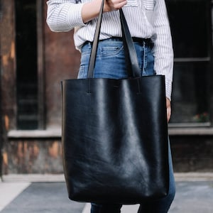 May include: A black leather tote bag with long straps. The bag is being held by a person wearing a striped shirt and blue jeans.