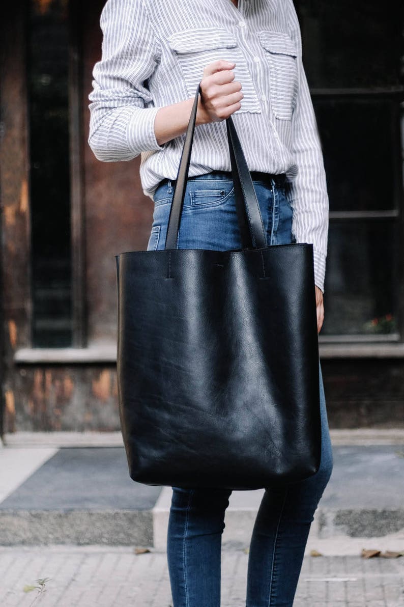 May include: A black leather tote bag with long straps. The bag is being held by a person wearing a striped shirt and blue jeans.