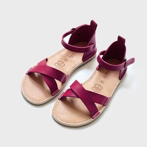 May include: A pair of burgundy leather sandals with criss-cross straps and an ankle strap with a buckle fastening.
