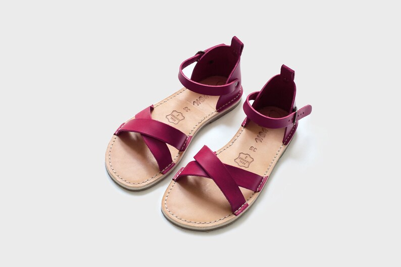 May include: A pair of burgundy leather sandals with criss-cross straps and an ankle strap with a buckle fastening.