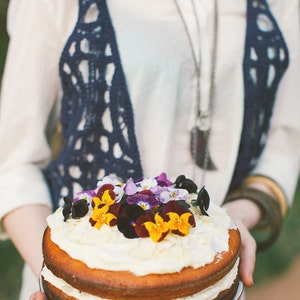 May include: A two-tiered cake decorated with a white frosting and a variety of colorful flowers. The cake is being held by a person wearing a white shirt and a blue crocheted vest.