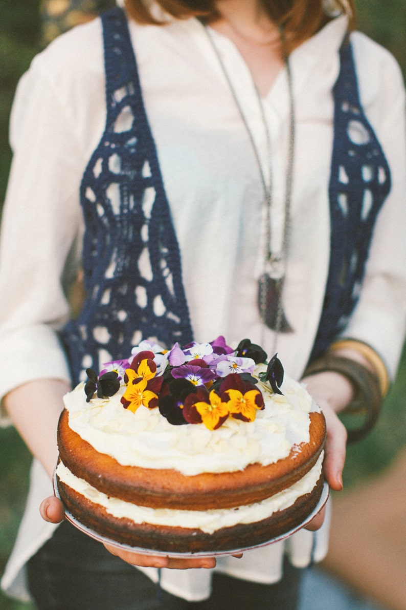 May include: A two-tiered cake decorated with a white frosting and a variety of colorful flowers. The cake is being held by a person wearing a white shirt and a blue crocheted vest.