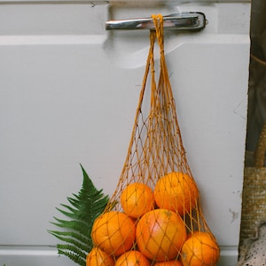 May include: A string bag filled with oranges hangs from a door handle. The bag is made of a fine mesh and the oranges are a bright orange color. The bag is hanging on a white door with a silver handle.