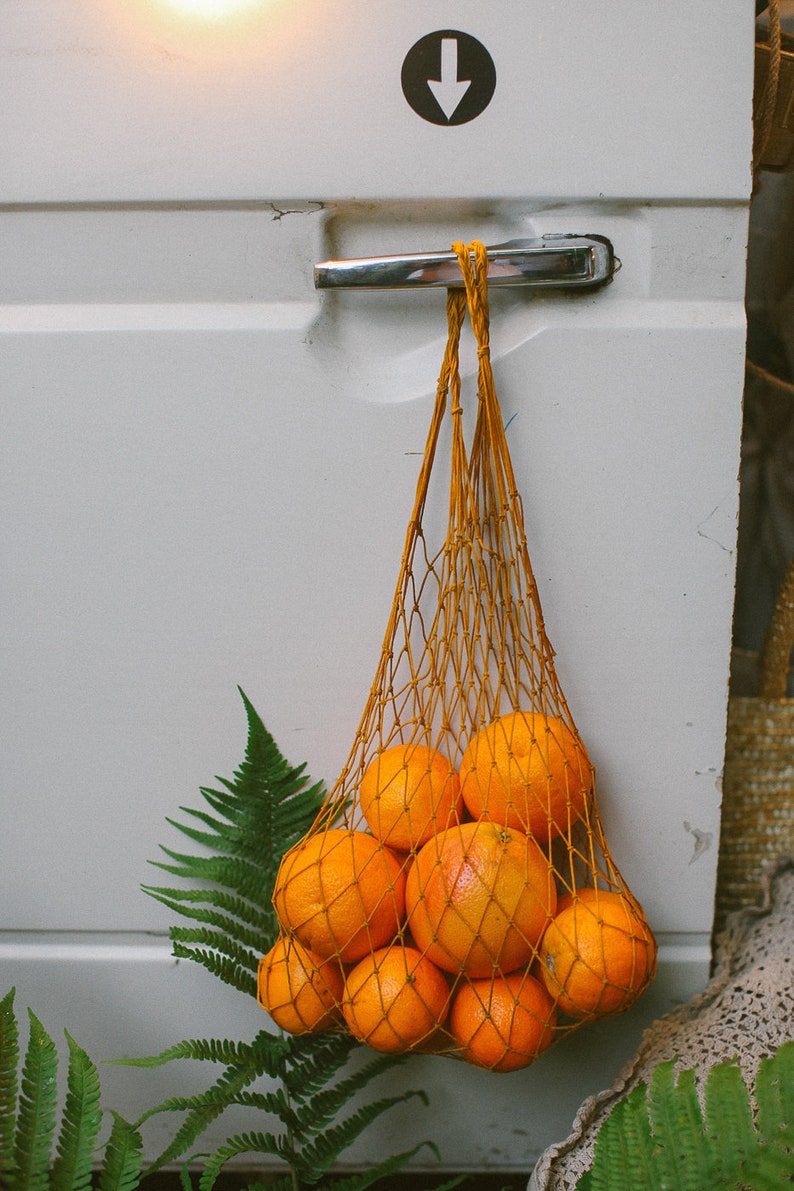 May include: A string bag filled with oranges hangs from a door handle. The bag is made of a fine mesh and the oranges are a bright orange color. The bag is hanging on a white door with a silver handle.
