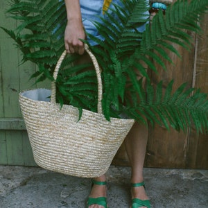 May include: A woman is holding a woven straw tote bag filled with green ferns. She is wearing a yellow top, denim shorts, and green sandals.