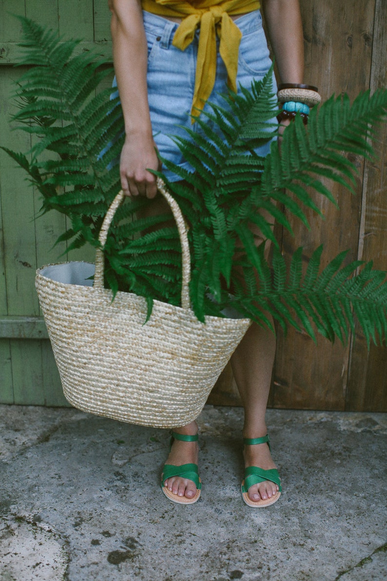 May include: A woman is holding a woven straw tote bag filled with green ferns. She is wearing a yellow top, denim shorts, and green sandals.