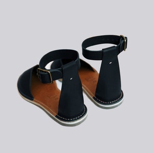 May include: A pair of black leather sandals with ankle straps and a brown leather insole. The sandals have a white stitching detail around the sole.