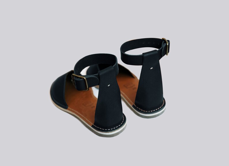 May include: A pair of black leather sandals with ankle straps and a brown leather insole. The sandals have a white stitching detail around the sole.