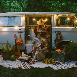 May include: Two people sitting in a white van with fairy lights, a guitar, and a watermelon. They are surrounded by sunflowers and a picnic blanket.