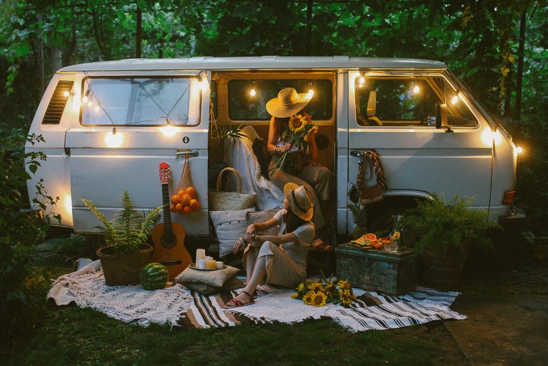 May include: Two people sitting in a white van with fairy lights, a guitar, and a watermelon. They are surrounded by sunflowers and a picnic blanket.