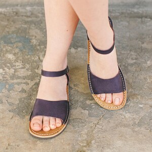 May include: A pair of dark blue leather sandals with a simple design and a brown sole. The sandals are being worn on someone's feet.