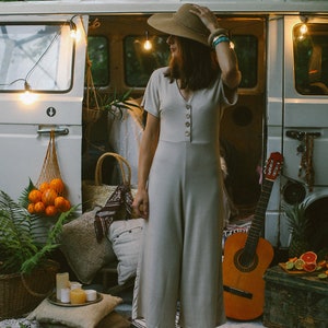 May include: A woman wearing a beige jumpsuit and a wide-brimmed hat stands in front of a vintage van. The van is decorated with string lights and has a guitar leaning against it. The woman is wearing blue sandals and has a turquoise bracelet on her wrist.