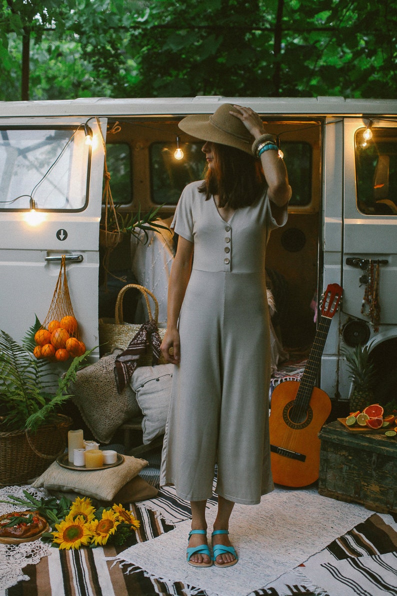 May include: A woman wearing a beige jumpsuit and a wide-brimmed hat stands in front of a vintage van. The van is decorated with string lights and has a guitar leaning against it. The woman is wearing blue sandals and has a turquoise bracelet on her wrist.