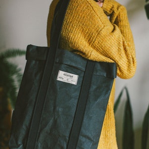 May include: A black tote bag with black straps and a white label that says "recycled". The bag is made of a textured material and is being held by a person wearing a yellow sweater.