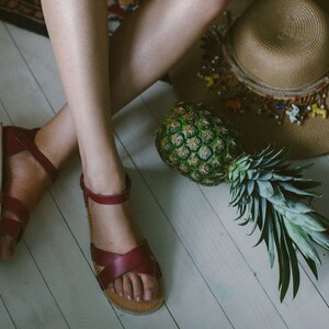 May include: A woman wearing burgundy leather sandals with a pineapple and a straw hat on the floor.