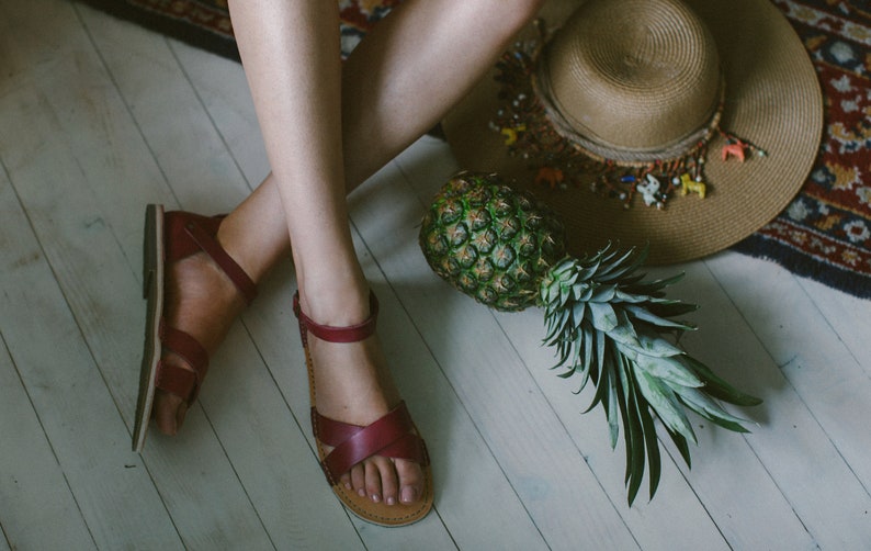 May include: A woman wearing burgundy leather sandals with a pineapple and a straw hat on the floor.