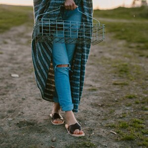 May include: A person wearing blue jeans and a striped blanket walks down a dirt path while carrying a wire basket.